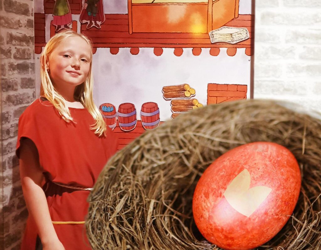Mädchen steht vor einer Wand im Museum. Im Vordergrund ist ein Nest mit einem Osterei.
